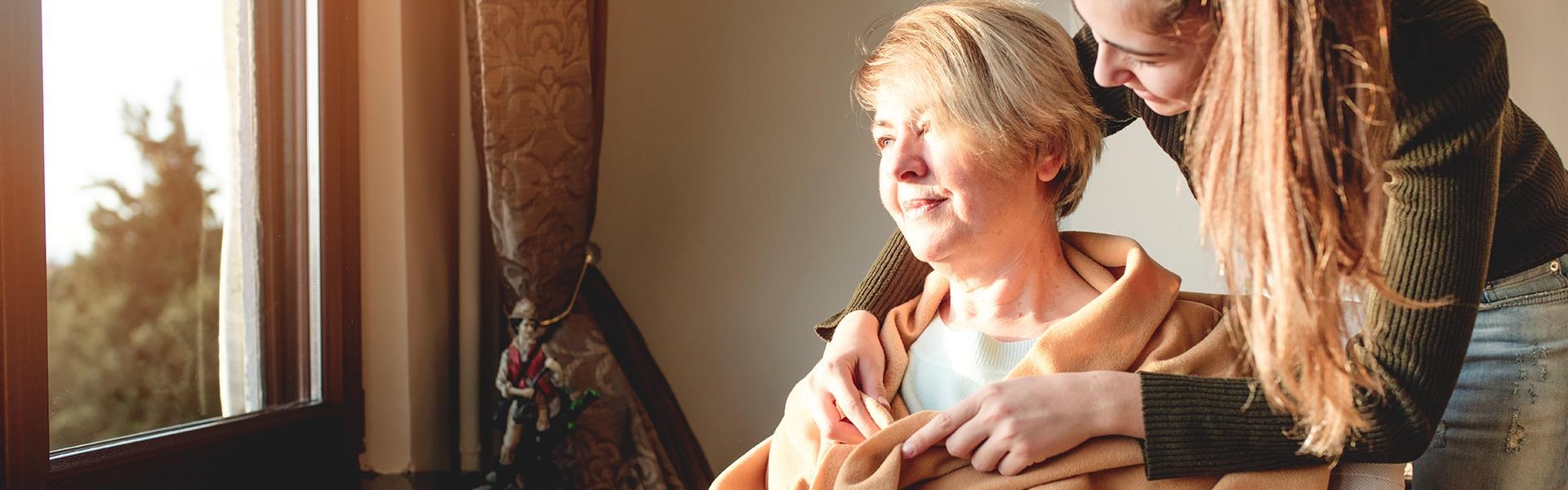 Junge erwachsene Person legt der älteren Person liebevoll eine Decke um, beide lächeln, Sonnenlicht fällt durch das Fenster