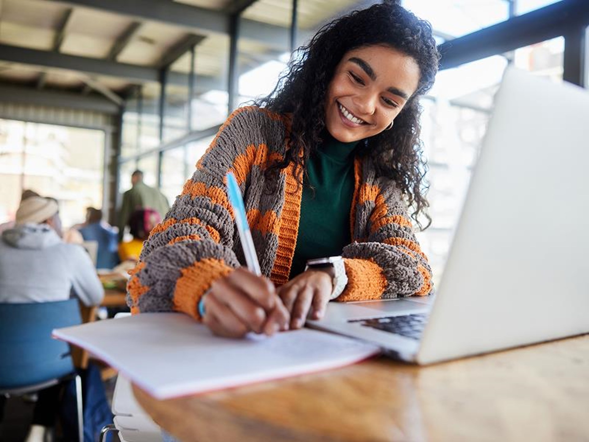 Lächelnde Person schreibt Notizen auf ein Blatt Papier, während sie an einem Laptop in einem hellen, modernen Arbeitsraum sitzt.