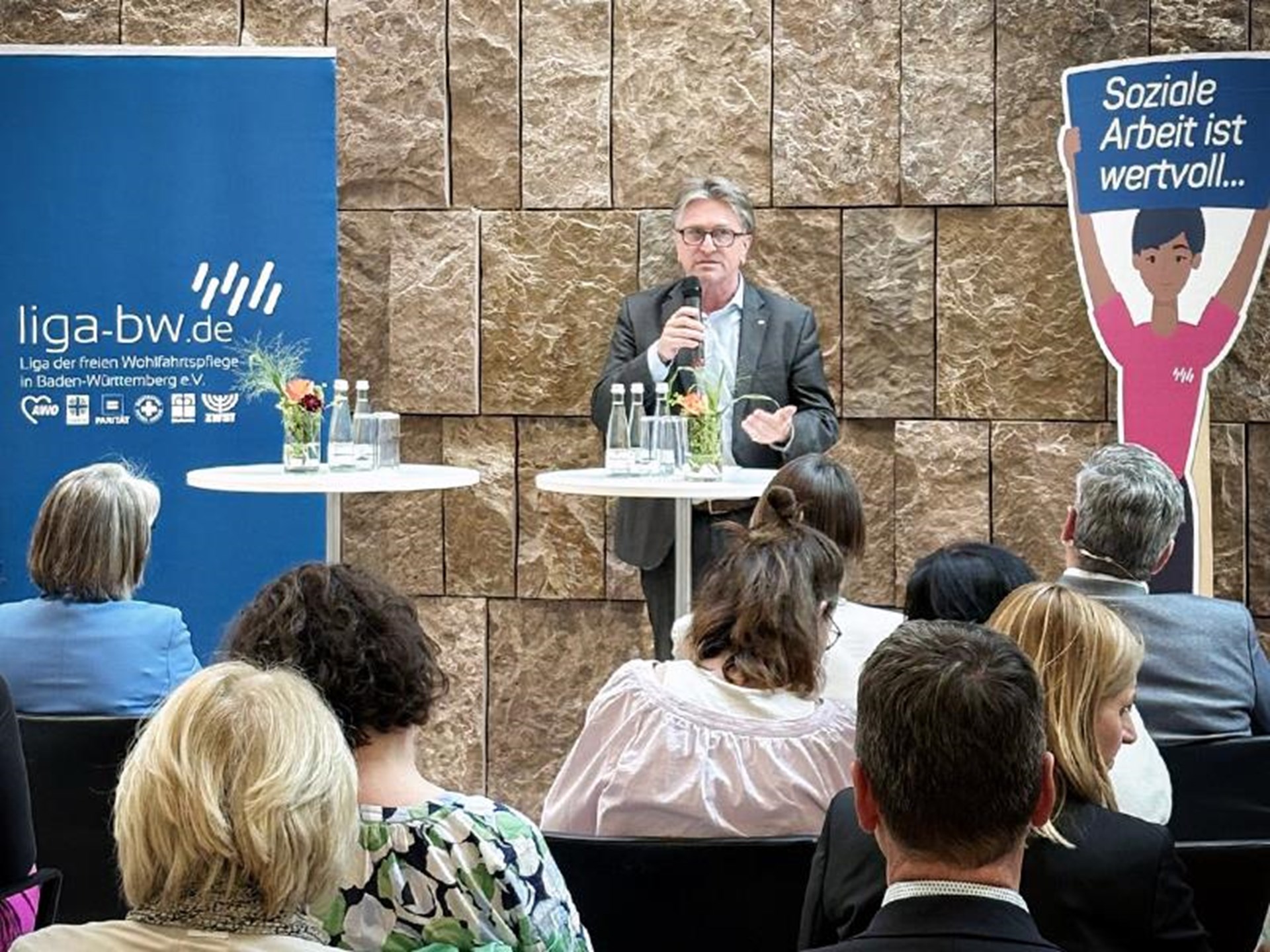 Redner spricht bei einer Veranstaltung der Liga der freien Wohlfahrtspflege, im Hintergrund Banner und Pappfigur mit Aufschrift 'Soziale Arbeit ist wertvoll'