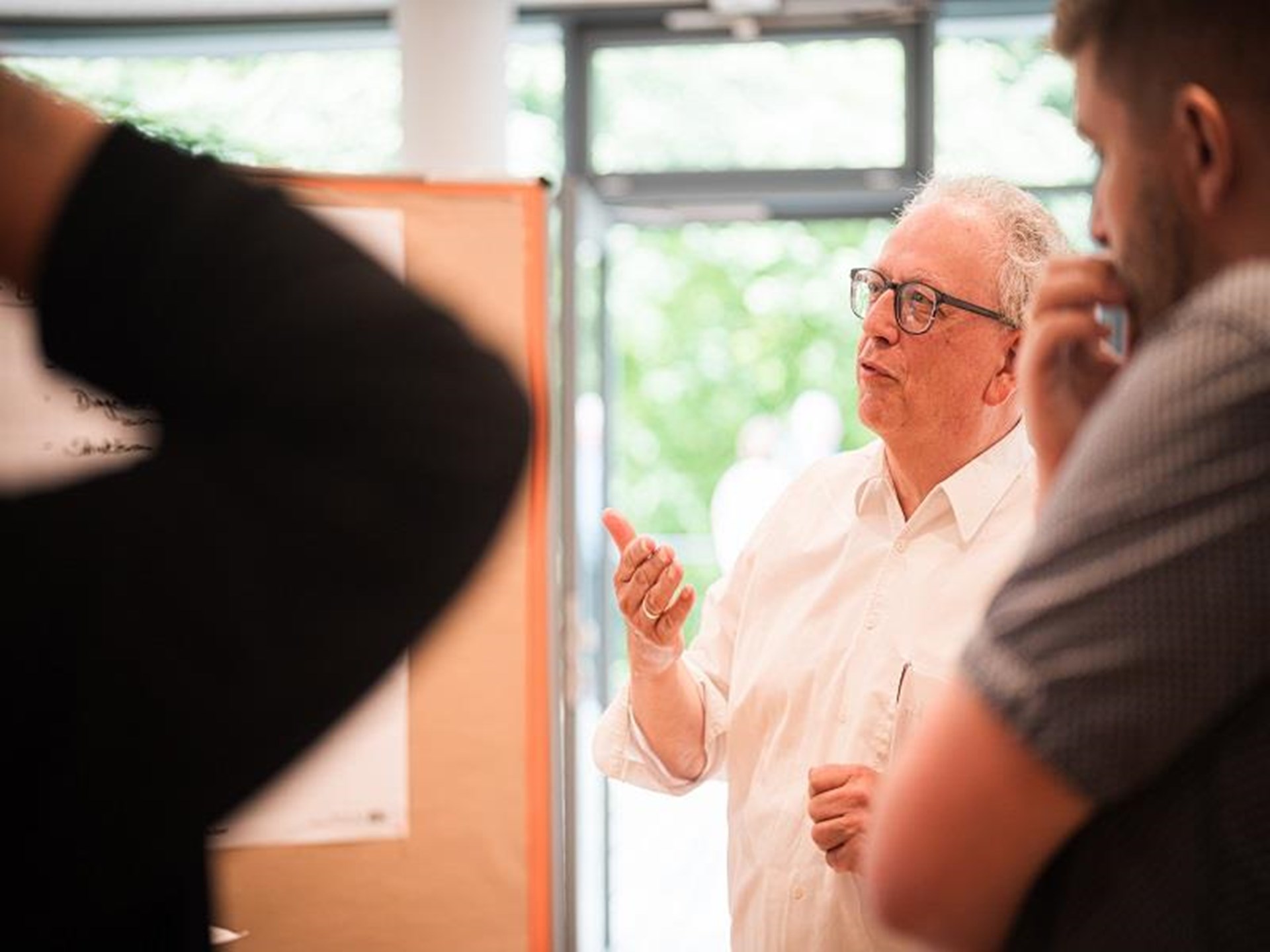 Ein Mann mit lockigen kurzen weißen Haaren und Brille steht vor zwei Erwachsenen Menschen hebt seine rechte Hand und erzählt etwas.